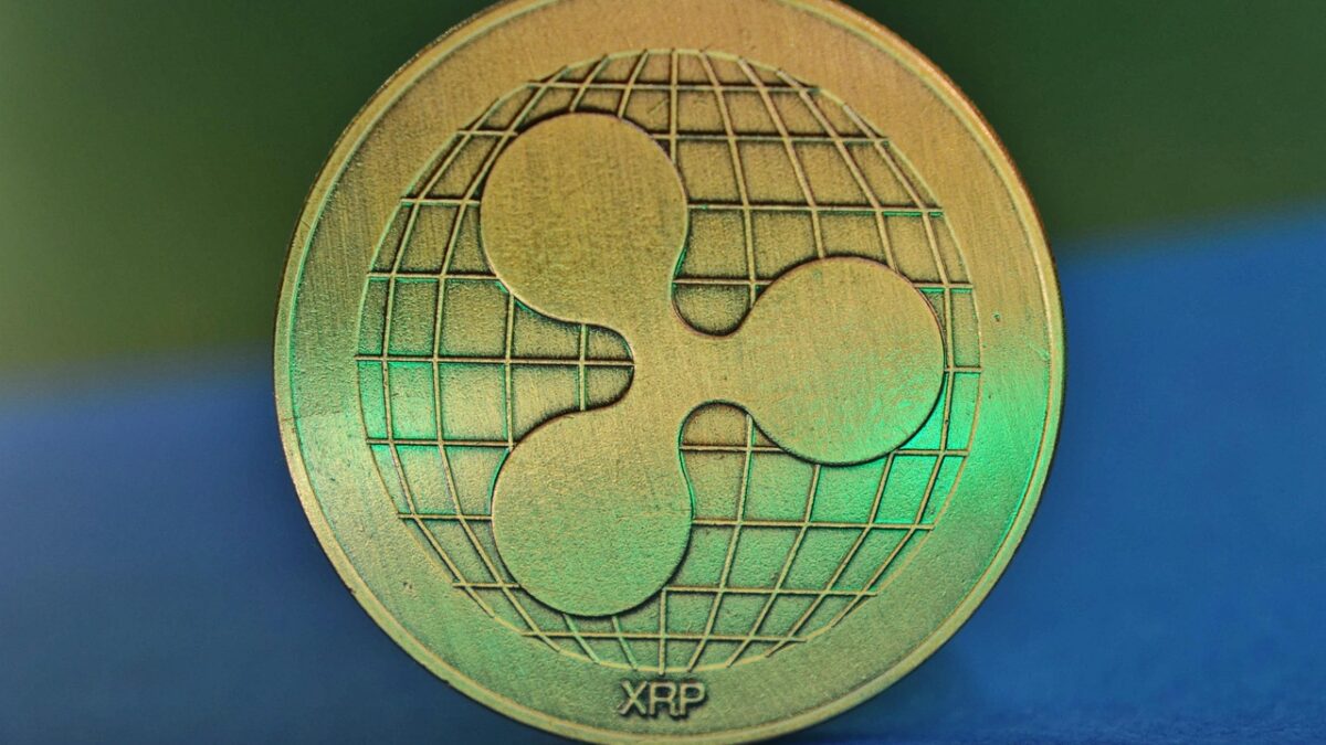 Gros plan d'une pièce dorée en relief marquée du logo Ripple, un globe quadrillé avec un symbole en trois formes, et de l'inscription « XRP », sur fond dégradé vert et bleu.