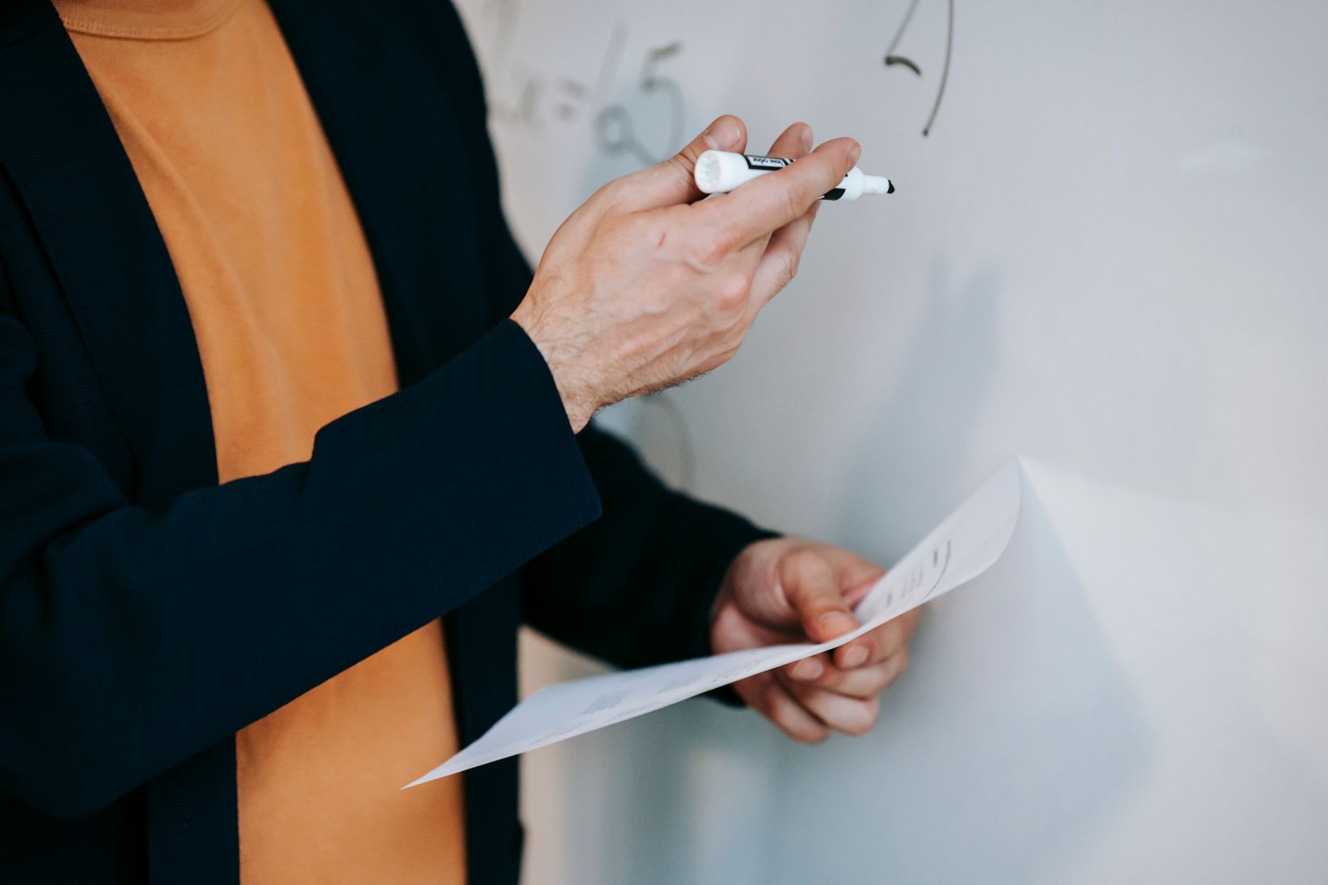 Gros plan d'un enseignant expliquant des concepts mathématiques sur un tableau blanc tout en tenant des notes.